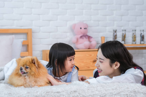 Menina asiática bonita e uma menina pequena estão olhando um para o outro com sorrisos, enquanto um cão está deitado ao lado deles na cama no quarto - Foto, Imagem