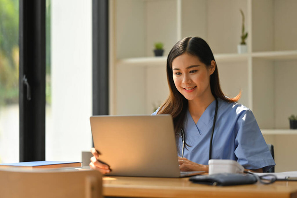 Portré mosolygós női orvos visel köpeny segítségével laptop a modern orvosi rendelőben. - Fotó, kép