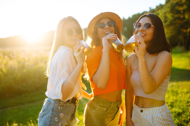 Three woman resting in nature in the picnic drinking beer, cheers. Vacation, picnic, friendship or holliday concept. - Foto, Imagen