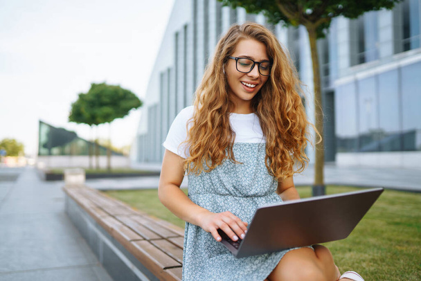 Smiling female freelancer sits on city street with laptop and wireless headphones calls on the laptop. Young woman with laptop works outdoors. Concept of education, online, business - Zdjęcie, obraz