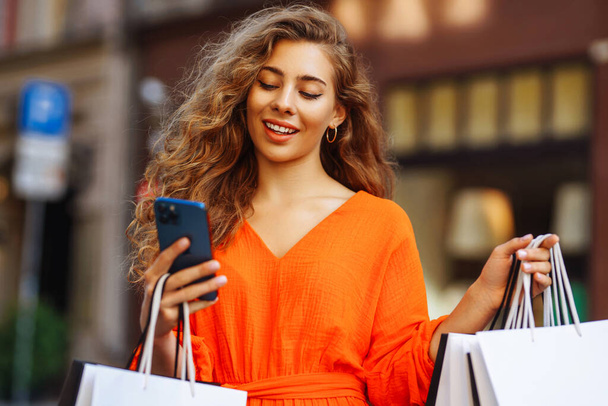 Shopping woman. Young woman with shopping bags in city with phone makes online purchases. Smiling tourist enjoys sunny weather while walking through streets of Europe. Online shopping concept. - Foto, immagini