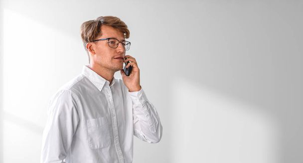 Businessman in eyeglasses calling on the phone, portrait in white shirt on empty copy space shadow wall. Concept of business network, online communication and conference - Zdjęcie, obraz