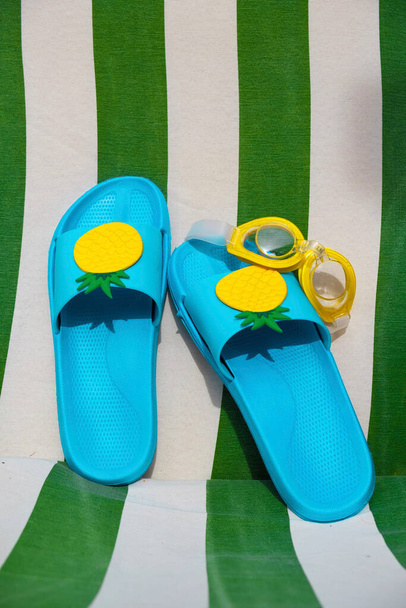 Blauwe poolsandalen en een gele poolbril op een groen-wit gestreepte stof. Zomer concept. Verticaal beeld. - Foto, afbeelding