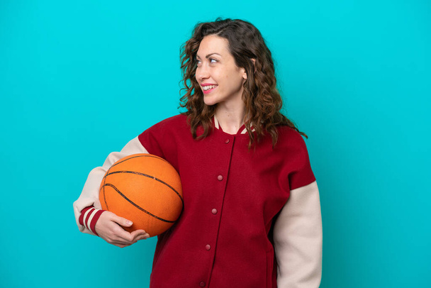 Fiatal kaukázusi kosárlabda játékos nő elszigetelt kék háttér néz oldalra, és mosolygós - Fotó, kép