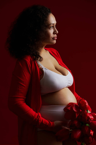 Μόδα πορτρέτο μιας όμορφης πολυεθνικής εγκύου, που φοράει λευκά εσώρουχα και κόκκινο ξεκούμπωτο πουκάμισο, κρατώντας ένα μάτσο κόκκινες τουλίπες, ποζάροντας γυμνή κοιλιά, στέκεται πλάι σε ένα σκούρο κόκκινο φόντο - Φωτογραφία, εικόνα