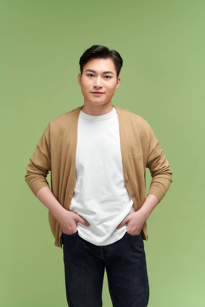 Photo de joyeux jeune asiatique gars mains poches isolé sur fond vert - Photo, image