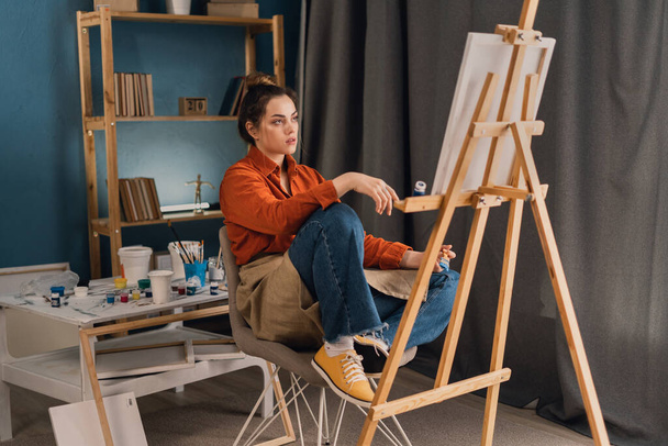 young latin woman artist sits in a home studio on a chair holds a palette with paints in her hands looks at the canvas and thinks. vacation and hobby concept. - Photo, Image