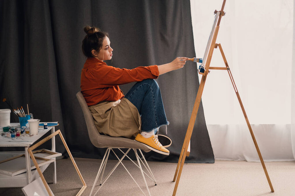 A beautiful Brazilian woman artist with paint on her hands works on painting a picture on canvas while sitting in front of her, on a stool. home studio. hobby and vacation concept. - Photo, Image