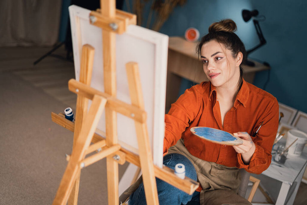 Yan bakışlı, gündelik giyinen etnik bir kadın sandalyede oturur, renkli boyalar çizerek tuvale çizer ve küçük odada fırçalar. Tuval üzerinde boya yapar. - Fotoğraf, Görsel