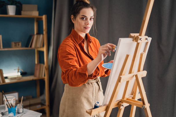 Творча латинська художниця, яка стоїть на новій картині. Щаслива молода леді, яка малює на полотні у своїй художній студії. Весела молода художниця працює в її домашній майстерні
. - Фото, зображення