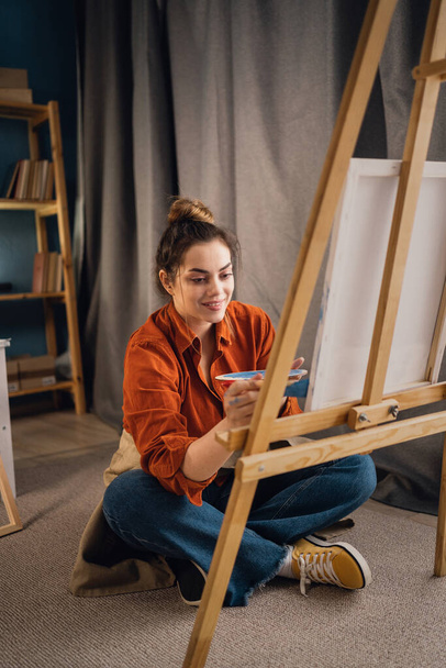 Young brazilian woman in art studio, learning to paint paintings on canvas, trying to mix different watercolors on cardboard. Concentrated woman with dark hair, casually dressed, painting. - Foto, Bild