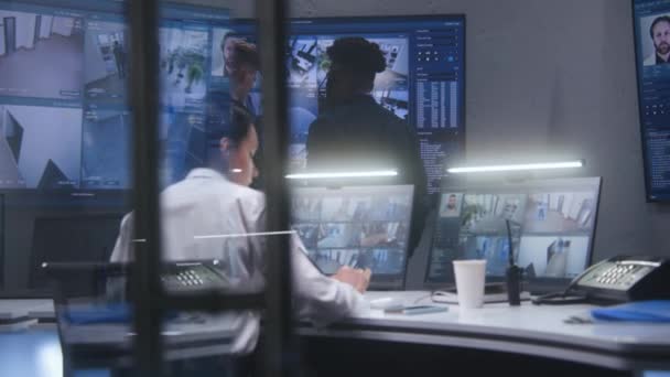 Biztonsági alkalmazottak figyelik a térfigyelő kamerák felvételeit AI arcszkenneléssel a rendőrségi megfigyelőközpontban. Az afro-amerikai operátor fülhallgatóban ül a munkahelyén. Képernyők a falon biztonsági kamerák felvételeivel. - Felvétel, videó