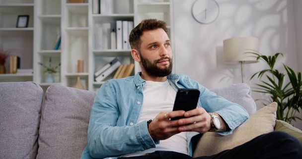 Portrait of attractive young man sitting on the sofa using phone smile in the modern apartment texting message content on smartphone social media applications online scrolling tapping technology. - Fotó, kép