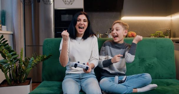 Mother and son playing video game using controllers when they spend joint leisure at home. Family celebrating victory in video game and gives five. Entertainment and modern lifestyle concept. - Photo, Image