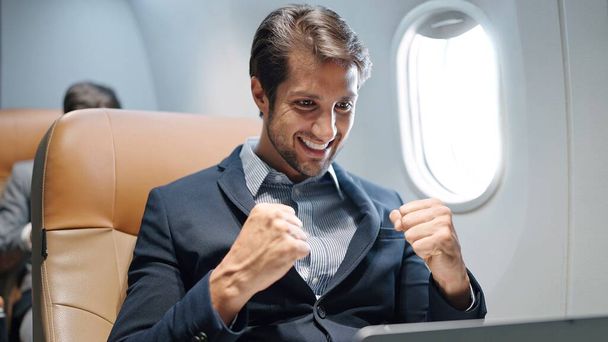 Handsome Indian or latin businessman wearing suit working on laptop hands up for celebration a good news of work sitting in seat on the plane at airplane - Foto, Bild