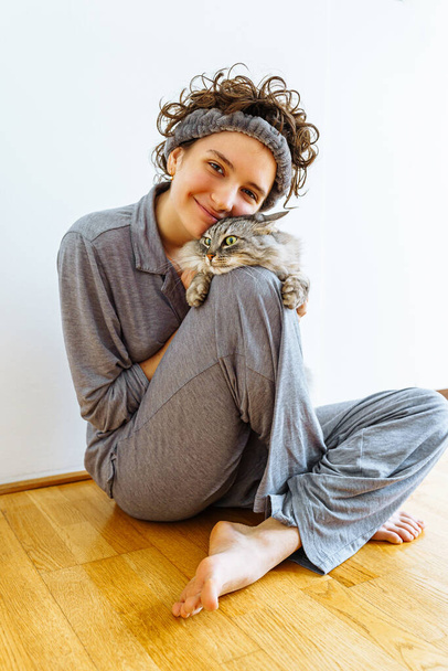 Jeune fille embrassant le chat bien-aimé du Maine Coon assis sur le sol à la maison, portant un pyjama et un bandeau, concept d'amour et de soins pour les animaux de compagnie - Photo, image