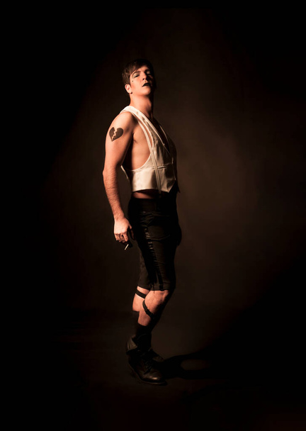 Master of ceremonies of a cabaret of the 20's, looking defiantly at the camera with a white vest and black shorts. In one hand a cigarette and black lips painted. Studio photo - Zdjęcie, obraz