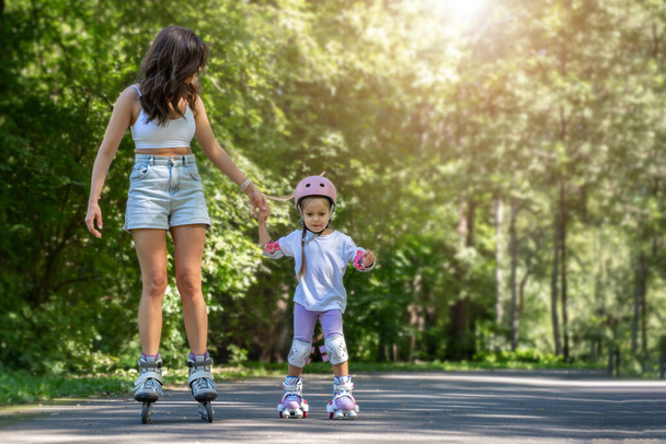 Mother teaching child daughter to skate on inline skates rollers in public park in summer. Family leisure outdoor sport activity game. Copy negative space - Photo, Image