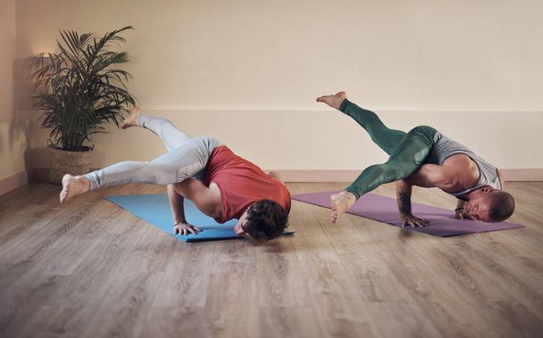 Wir stoßen an unsere Grenzen. Aufnahme in voller Länge von zwei jungen Männern, die während einer gemeinsamen Yoga-Stunde im Innenraum eine ausgedehnte seitliche Krähenpose halten - Foto, Bild