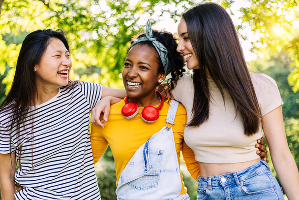 3人の多人種の女性の若いグループが屋外で一緒に楽しんでいます。市内の公園を歩いている100万人の女性の友人の笑顔の肖像画。友情と若者のライフスタイル. - 写真・画像