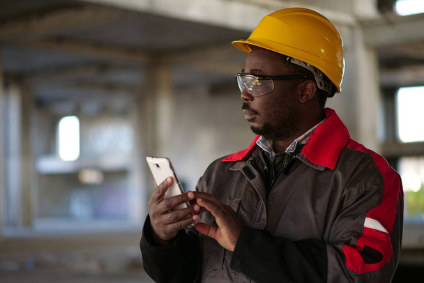 スマートフォンを備えた黄色のハード帽子のアフリカ系アメリカ人労働者は、建設現場に立っています。スマートフォンを手に持つ労働者 - 写真・画像