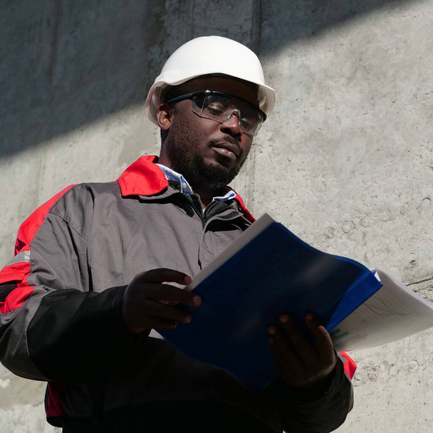 Afrykański amerykański robotnik stoi na placu budowy i wygląda jak papier roboczy - Zdjęcie, obraz