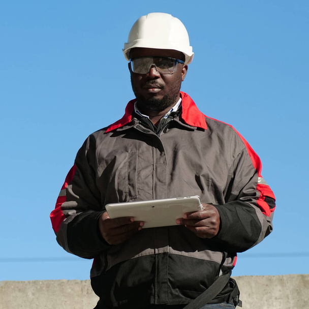 Afrikkalainen amerikkalainen työntekijä valkoisessa kypärässä ja suojalaseissa seisoo rakennustyömaalla, pitää käsissään tabletin tietokonetta ja katsoo kameraa - Valokuva, kuva