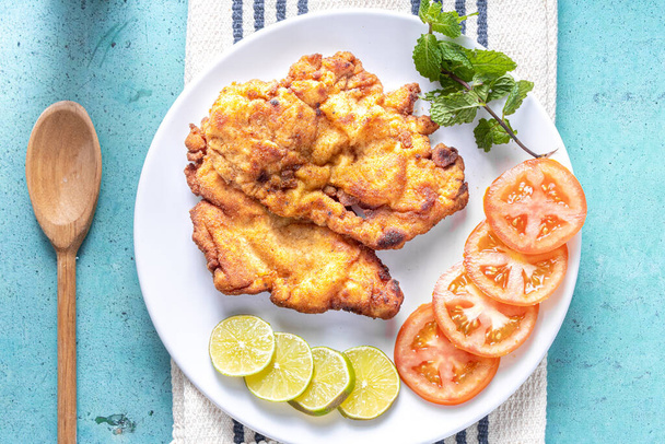 Delicious Milanese chicken (breaded chicken with breadcrumbs and egg), a typical Brazilian dish. On a white plate with tomatoes, lemon on striped white tablecloth and blue marble. Top view. - Photo, Image