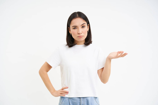 So what. Young asian woman shrugs shoulds, looks questioned at camera, cant understand, stands over white background. - Photo, Image