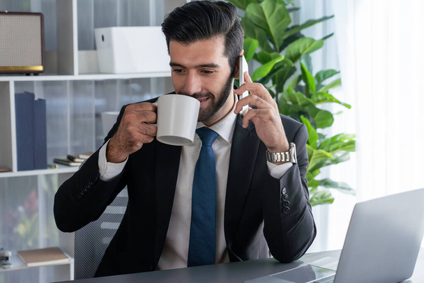 Üzletember dolgozik a modern irodai munkaterületen egy csésze kávéval a kezében, miközben válaszol a telefonhívásokra, értékesítési hívásokra vagy az alkalmazottak kezelésére. Fertőzött - Fotó, kép