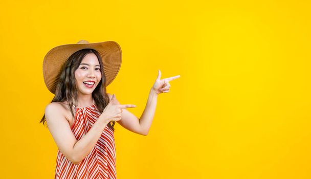 Estúdio retrato tiro de mulher asiática em vestido sexy com chapéu de palha sorrindo em felicidade apontando gesto como apresentador isolado no fundo amarelo para viagens e férias de verão - Foto, Imagem