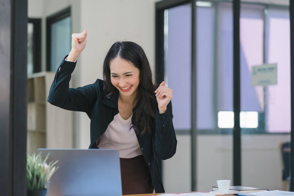 Bir iş kadını mutlu ve başarılı bir pozda dizüstü bilgisayara bakarken kollarını kaldırarak kutlama yapar. - Fotoğraf, Görsel