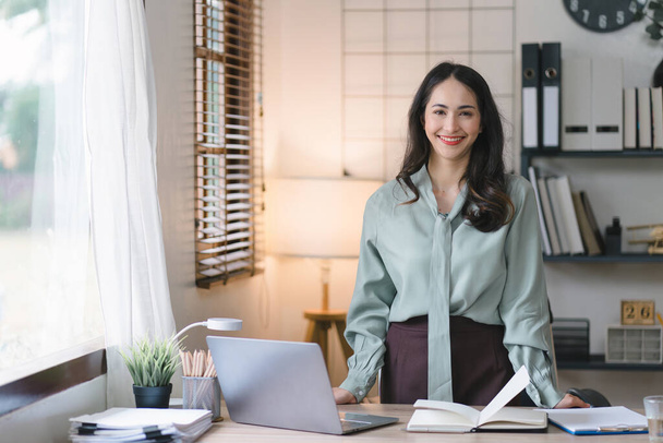 Imagem de uma jovem, alegre, feliz, positiva, fofa e bela mulher de negócios milenar asiática em pé dentro de casa em um escritório em casa, usando um computador portátil - Foto, Imagem