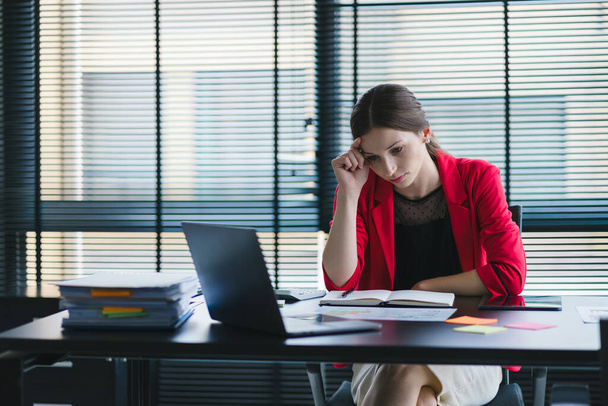 La femme d'affaires millénaire stressée et épuisée est vue assise à son bureau la main sur la tête, ce qui indique une dure journée de travail où elle est surchargée de travail - Photo, image