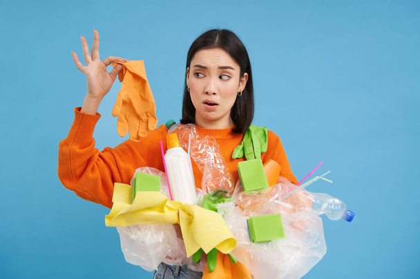 Young woman holds plastic garbage, looks at glove with shocked face, stinky waste, sorting litter for recycling, blue background. - Fotó, kép