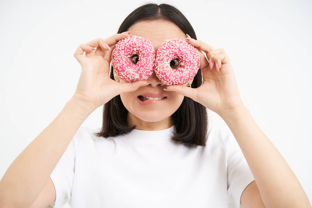 Image of funny young woman, makes glasses with two pink glazed doughnuts, looking through donnut holes and smiling, isolated on white background. - Photo, Image