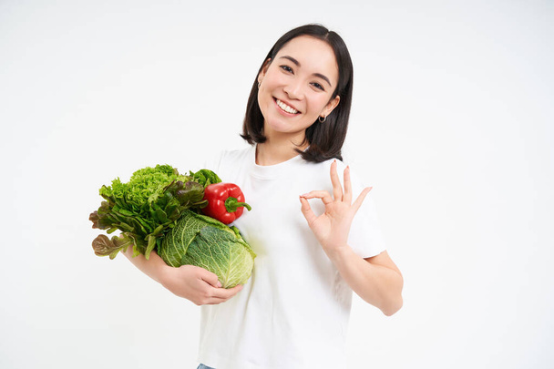 Улыбающаяся женщина, держа в руках овощи, показывает ладно, хорошо, знак, изолированный на белом фоне. - Фото, изображение