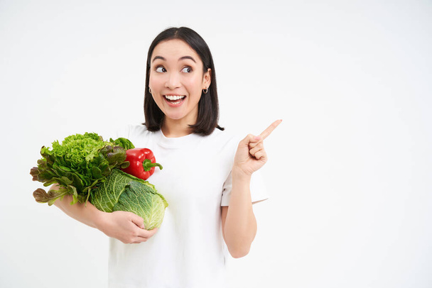 Mujer coreana entusiasta, sosteniendo racimo de verduras, señalando el dedo a la derecha en la pancarta, mostrando publicidad promocional, fondo blanco. - Foto, imagen