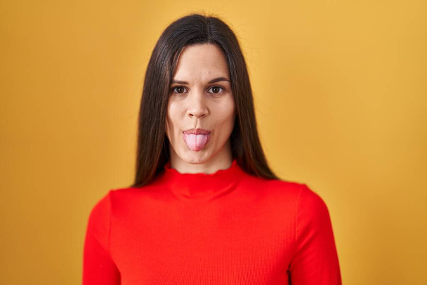 Young hispanic woman standing over yellow background sticking tongue out happy with funny expression. emotion concept.  - Zdjęcie, obraz