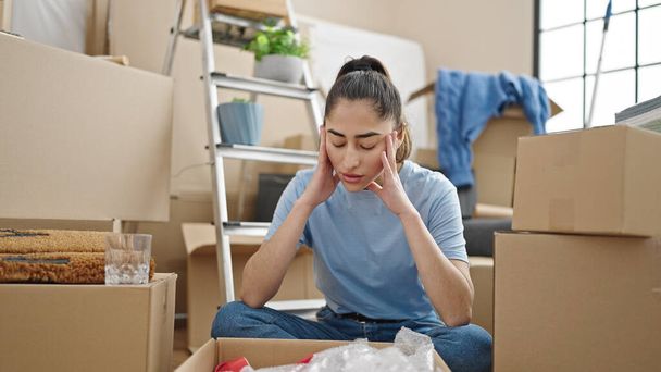Young beautiful hispanic woman unpacking cardboard box with serious expression at new home - Photo, Image