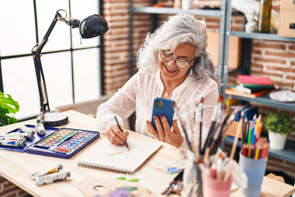 Middle age woman artist drawing on notebook using smartphone at art studio - Fotó, kép