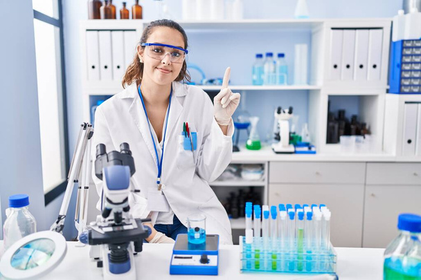 Young hispanic woman working at scientist laboratory showing and pointing up with finger number one while smiling confident and happy.  - Foto, Imagen