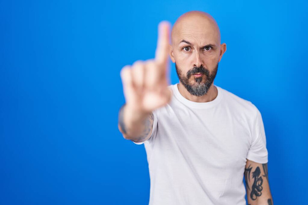 Spaanse man met tatoeages over blauwe achtergrond wijzend met vinger omhoog en boze uitdrukking, zonder gebaar  - Foto, afbeelding