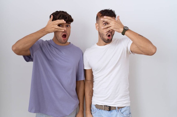 Homosexuální homosexuální homosexuální pár stojící nad bílým pozadím vykukující v šoku a zakrývající obličej a oči rukou, dívající se skrz prsty s rozpačitým výrazem.  - Fotografie, Obrázek