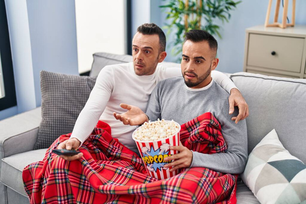 Homosexual couple eating popcorn watching tv clueless and confused expression. doubt concept.  - Фото, изображение