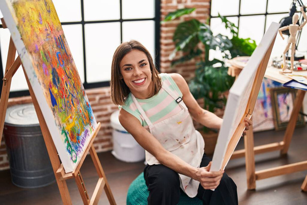 Νεαρή όμορφη Ισπανίδα καλλιτέχνης χαμογελώντας αυτοπεποίθηση κρατώντας κλήρωση στο στούντιο τέχνης - Φωτογραφία, εικόνα
