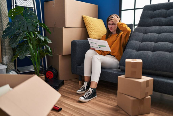 Молодая брюнетка, сидящая на диване в новом доме, смотрит на документы, напряженные и разочарованные рукой об голову, удивленное и сердитым лицом  - Фото, изображение