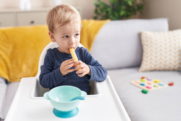 Αξιολάτρευτο ξανθό νήπιο κάθεται σε καρεκλάκι τρώει σνακ στο σπίτι - Φωτογραφία, εικόνα