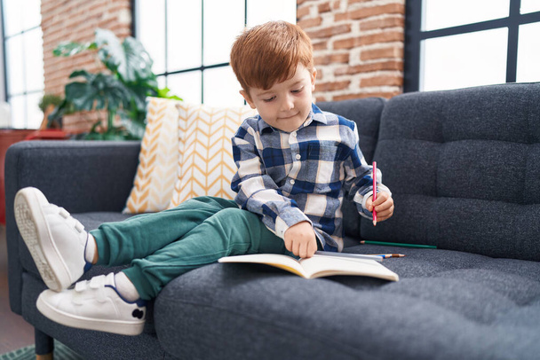 Adorable toddler drawing on notebook sitting on sofa at home - Fotó, kép