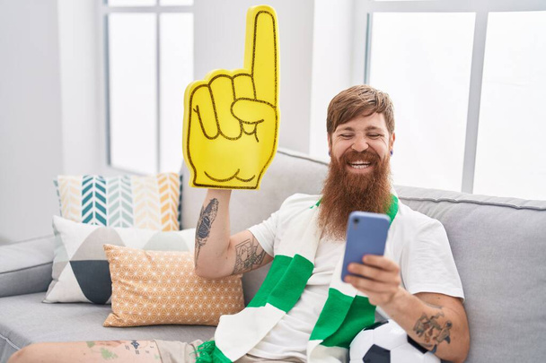 Fiatal vörös hajú férfi támogatja a focicsapat segítségével smartphone gazdaság nagy kéz otthon - Fotó, kép